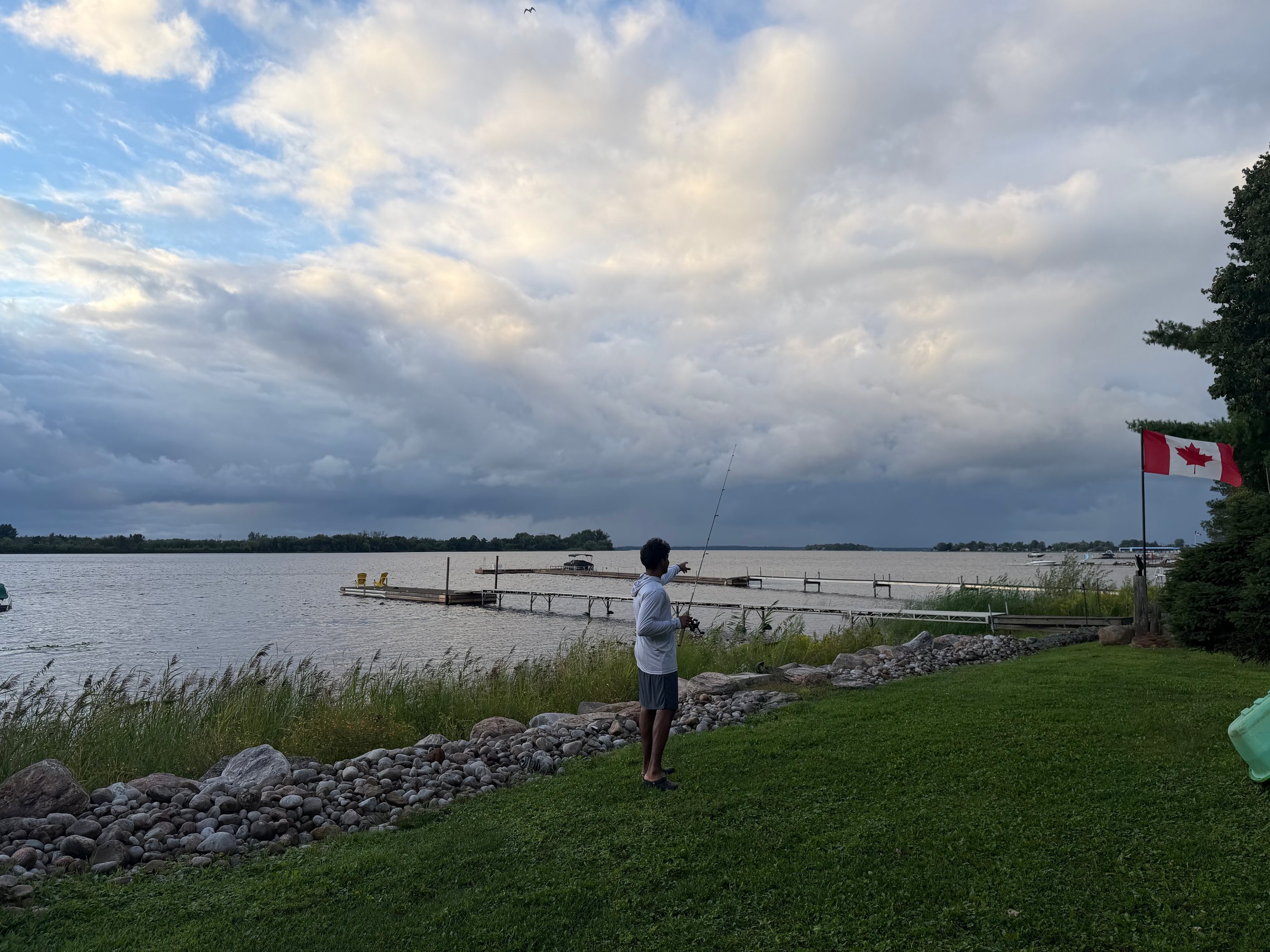Sheehan casting a fishing line from a rocky shore at dusk, pointing toward the far side of the lake, under a clearing storm sky.